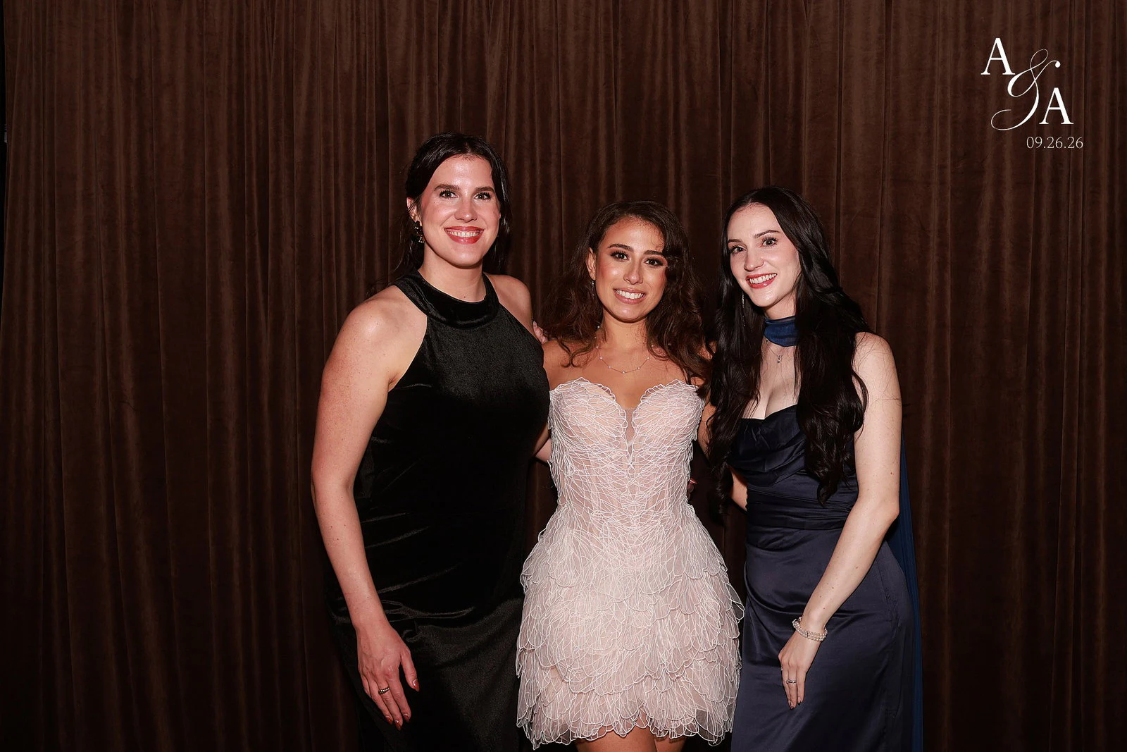 A woman in a pale feathered dress between two friends in black velvet and navy satin in front of the chocolate brown velvet at the Alyssa + Alex wedding