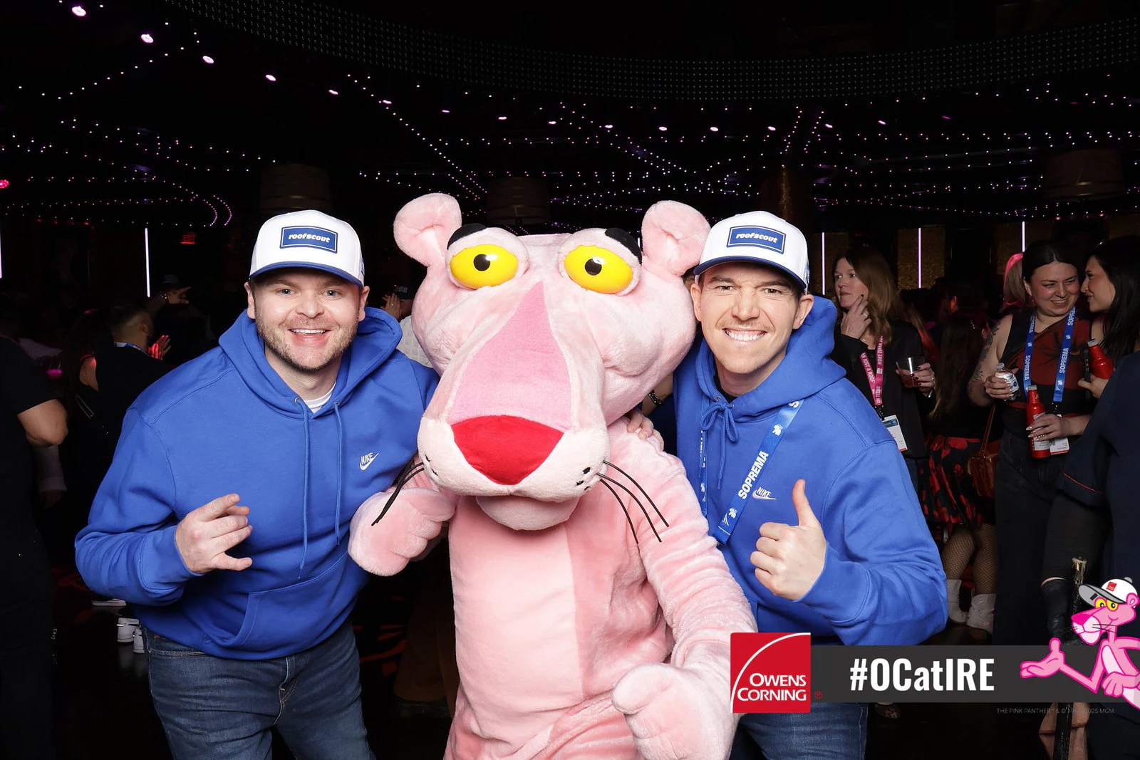 Two men in blue hoodies and white caps either side of a costumed pink panther mascot in a crowded club at Owens Corning at IRE