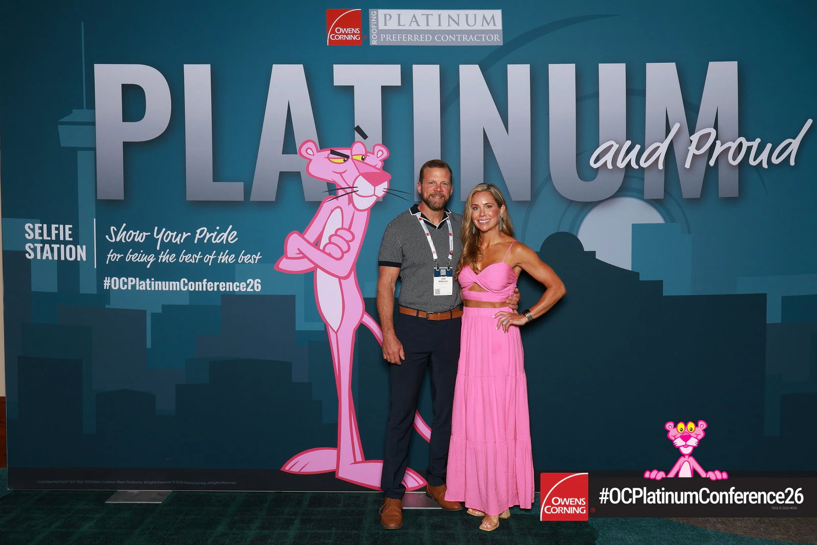 A man in a checked polo and a woman in a pink two-piece dress beside the pink cartoon panther on the teal printed wall at Platinum Conference 2026