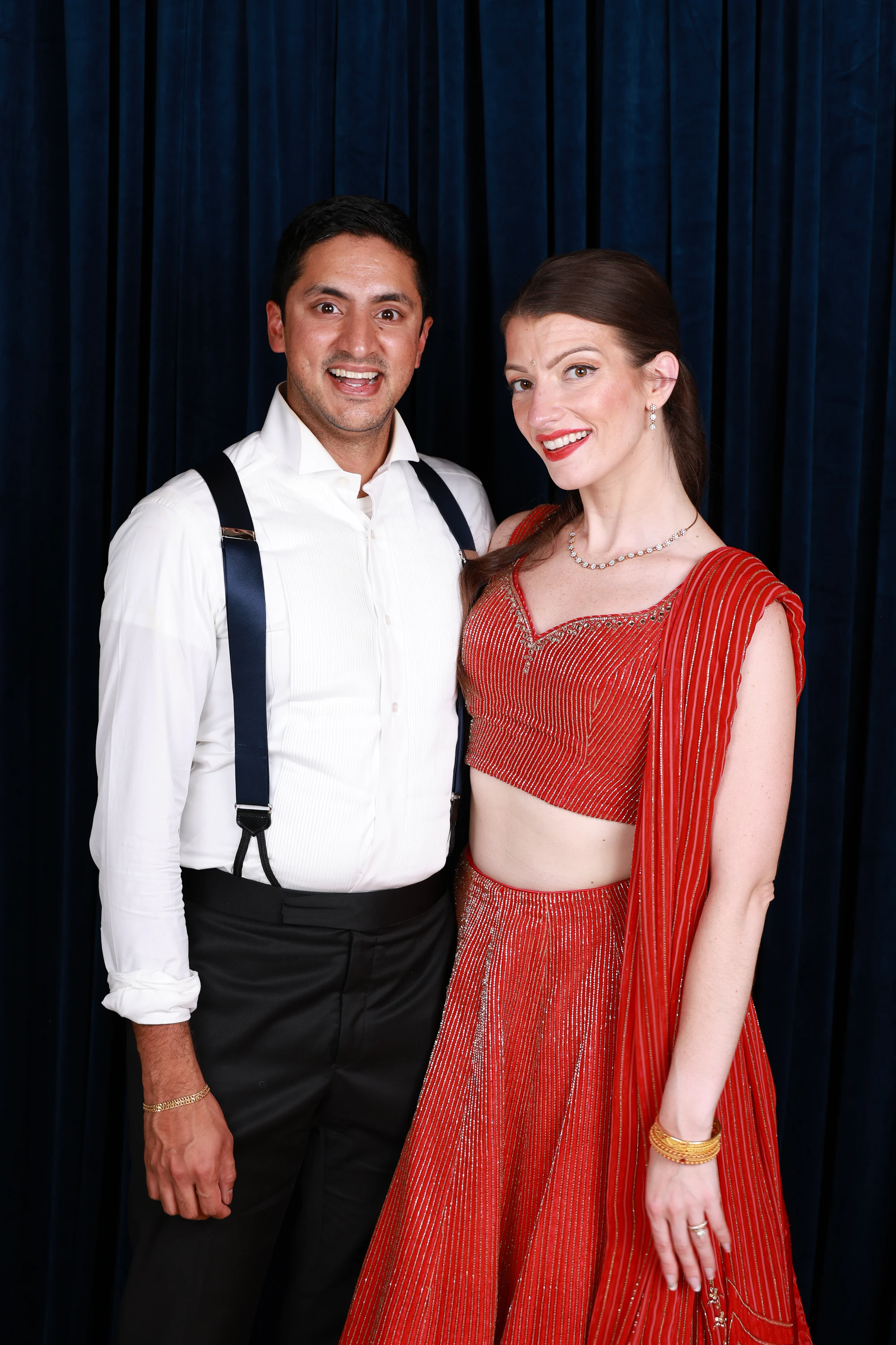 A couple in a red gown and suspenders, arm around, in front of the navy velvet at Ramya + Sreehari’s wedding