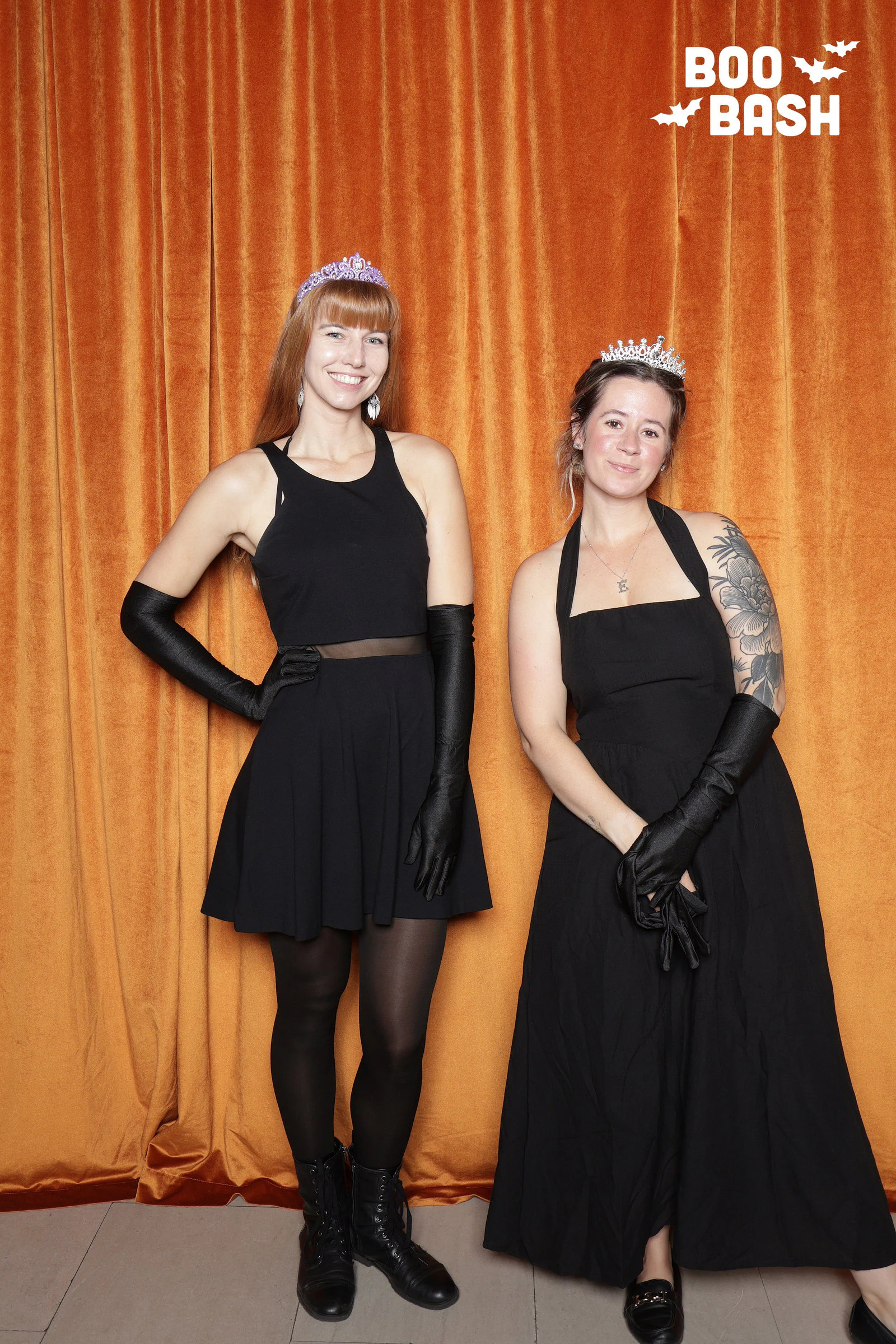 Two women in black dresses, tiaras and long black gloves in front of the orange velvet at Rocket Boo Bash