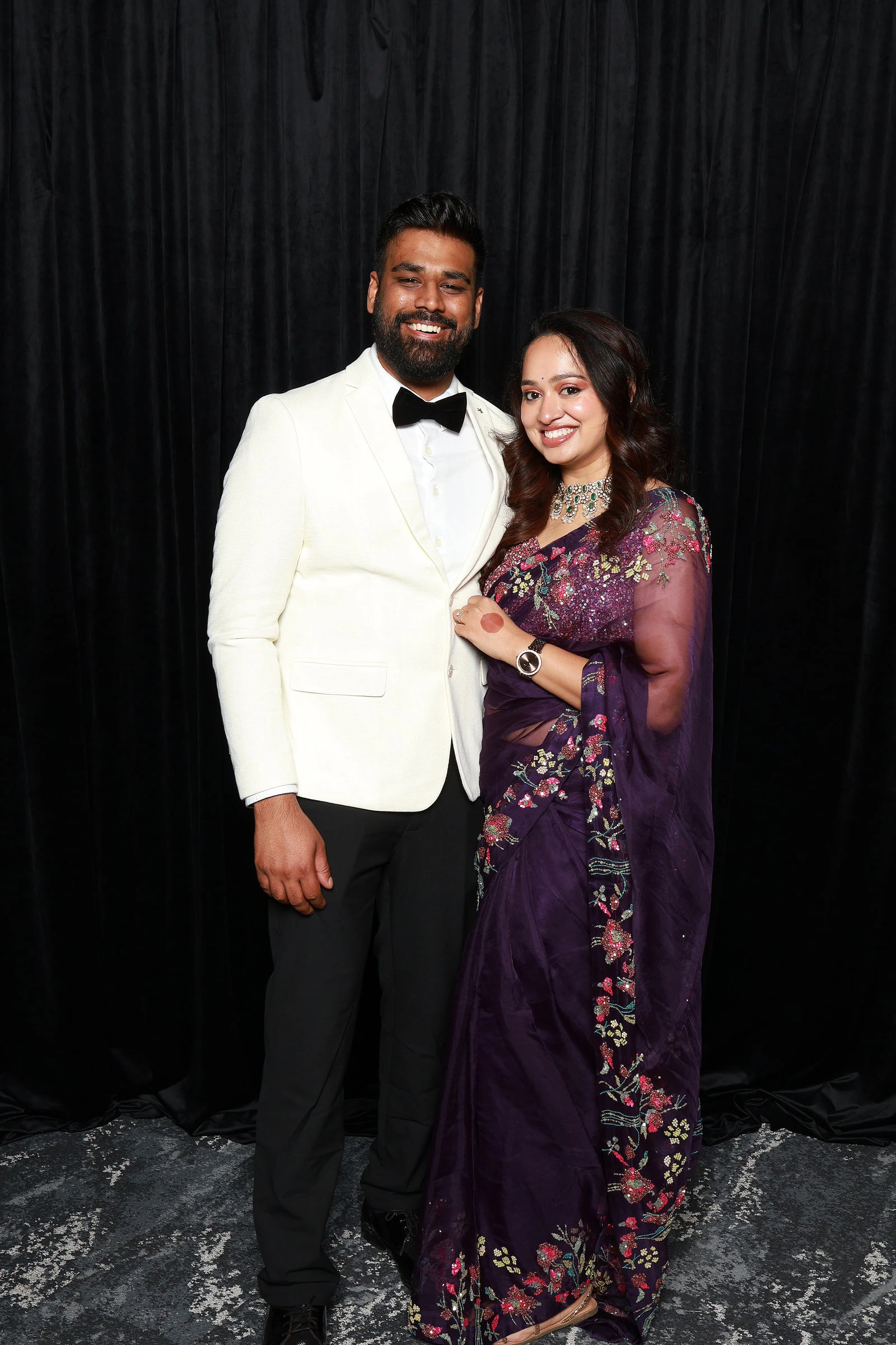 A man in a white dinner jacket and bow tie beside a woman in a purple embroidered sari in front of the black velvet at the Yamini + Hari wedding